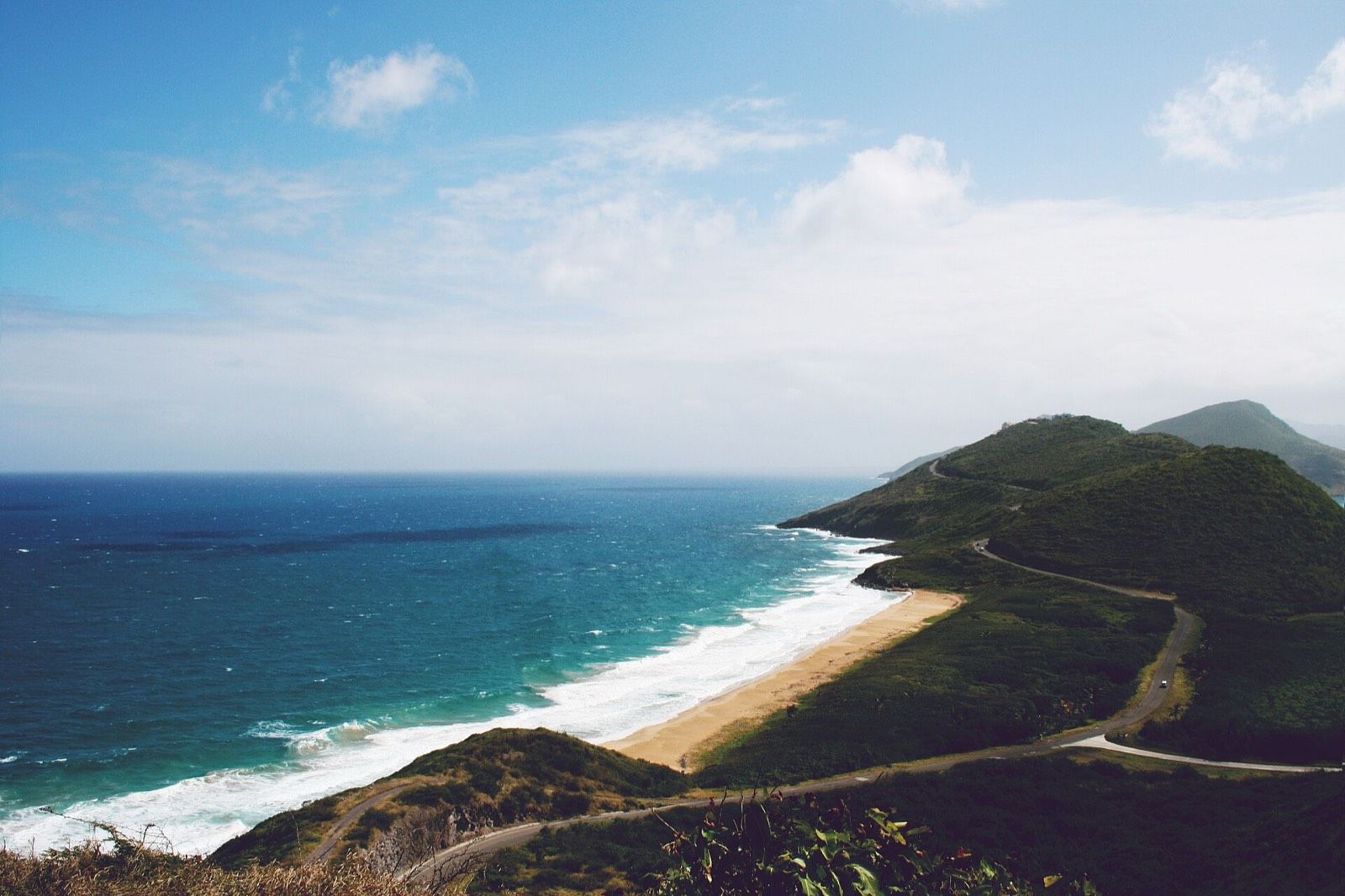 A long sandy beach on St. Kitts where Atlantic waves break, with a road winding over green hills beside it